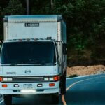 A white truck driving on a winding road surrounded by dense green forest plants.